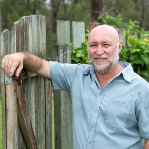 A man leans against a wooden fence, holding a green garden hose in a sunny outdoor setting.